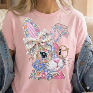 Puede incluir: Camiseta rosa claro con un diseño de conejo colorido. El conejo tiene orejas con estampado floral, un lazo y gafas redondas. El diseño incluye perlas y detalles florales, creando un aspecto caprichoso y decorativo.