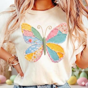 Könnte beinhalten: Cremefarbenes T-Shirt mit einem bunten Patchwork-Schmetterlingsdesign. Der Schmetterling hat Flügel in Rosa, Blau, Gelb und Grün mit floralen und gemusterten Details. Das Shirt ist ein lässiger Stil mit kurzen Ärmeln.