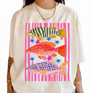 May include: Cream-colored t-shirt featuring a colorful fish design. The artwork includes three fish in yellow, orange, and pink, set against a pink and white striped background with blue and pink star accents. The shirt has short sleeves.