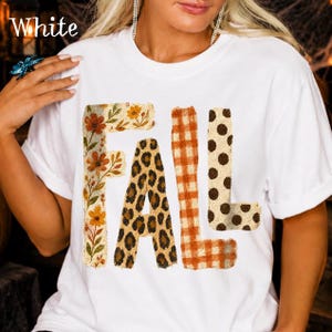 Może przedstawiać: Biały t-shirt z krótkim rękawem z napisem FALL wydrukowanym dużymi literami. Każda litera ma inny wzór: F ma nadruk kwiatowy, A nadruk w panterkę, L pomarańczową kratkę gingham, a druga litera L brązowe kropki.
