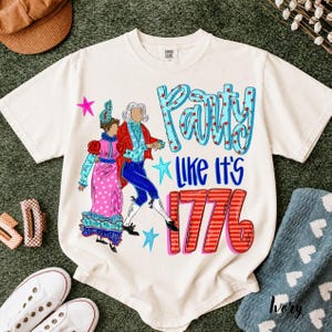 May include: An off-white short-sleeve t-shirt featuring a graphic of two figures in colonial-style clothing. The text reads Party Like It's 1776 in blue and red lettering. The design includes blue stars and pink accents on a light-colored fabric.