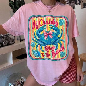 Puede incluir: Una camiseta rosa claro con el gráfico de un cangrejo azul que lleva un lazo a rayas rosas y blancas. El diseño incluye rayas verticales coloridas, conchas marinas y el texto "If Crabby Return To The Beach" en una fuente rosa y roja estilizada.
