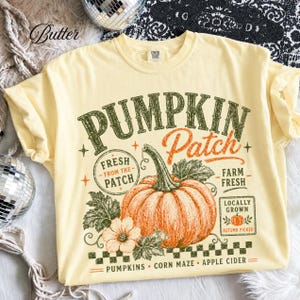 Pode incluir: Uma t-shirt amarelo-pálido com uma impressão gráfica de uma abóbora laranja grande com folhas e flores verdes. O texto diz PUMPKIN Patch, FRESH FROM THE PATCH, FARM FRESH, LOCALLY GROWN, AUTUMN PICKED, PUMPKINS, CORN MAZE e APPLE CIDER.