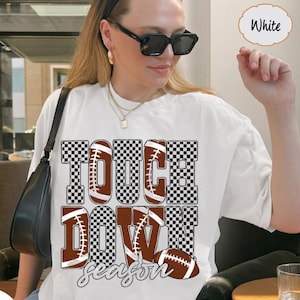 Könnte beinhalten: Ein weißes kurzärmeliges T-Shirt mit einem großen Grafikdruck mit dem Text TOUCH DOWN. Die Buchstaben sind mit einem braun-weißen Karomuster und braunen Football-Formen gefüllt. Eine schwarze Umhängetasche wird an der Seite getragen.
