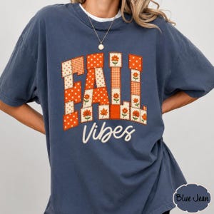 Könnte beinhalten: Ein schieferblaues Kurzarm-T-Shirt mit dem Text FALL VIBES. Das Wort FALL ist im Patchwork-Stil mit orangefarbenen und weißen Mustern wie Punkten, Vichy-Karos und Blumenmotiven bedruckt. Das Wort VIBES ist in weißer Schreibschrift gehalten.