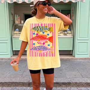 Puede incluir: Una camiseta de manga corta de color amarillo p&aacute;lido con un estampado gr&aacute;fico de tres peces apilados verticalmente. Los peces est&aacute;n representados en verde, naranja y rosa con patrones de manchas, sobre un fondo de rayas verticales rosas y estrellas azules.