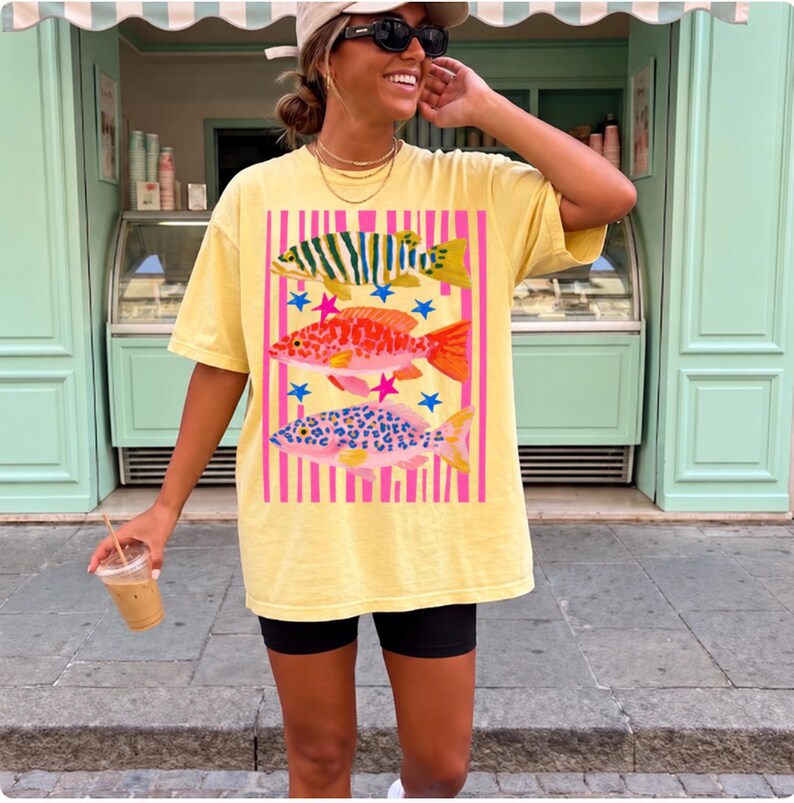 Puede incluir: Una camiseta de manga corta de color amarillo p&aacute;lido con un estampado gr&aacute;fico de tres peces apilados verticalmente. Los peces est&aacute;n representados en verde, naranja y rosa con patrones de manchas, sobre un fondo de rayas verticales rosas y estrellas azules.