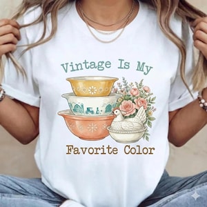May include: White t-shirt with the text "Vintage Is My Favorite Color." The graphic features a stack of vintage bowls in yellow, white, and orange, with floral and farm-themed designs. A bouquet of pink roses and a chicken in a bowl complete the design.
