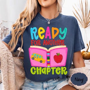 May include: A navy blue t-shirt featuring a graphic of an open pink book with a yellow school bus on the left page and a red apple on the right page. The text reads READY FOR ANOTHER CHAPTER in colorful, bold, playful lettering across the front.