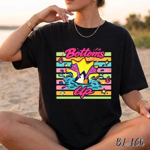 Puede incluir: Una camiseta negra con un estampado gráfico de un pato sumergiéndose en el agua, con sus patas palmeadas rosas visibles sobre la superficie. El diseño incluye rayas horizontales en amarillo, verde y rosa, con el texto Bottoms Up en letras rosas.