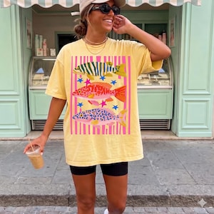 Puede incluir: Camiseta amarilla con un diseño de peces coloridos. El gráfico incluye tres peces con diferentes patrones y colores, sobre un fondo de rayas rosas y blancas con estrellas azules. La camiseta se usa con pantalones cortos negros.
