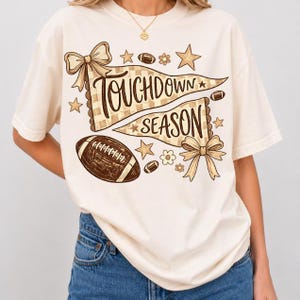 Könnte beinhalten: Ein cremefarbenes T-Shirt mit einem braunen Grafikdruck von zwei Wimpeln mit dem Text "TOUCHDOWN SEASON". Das Design enthält einen Football, Schleifen, Sterne und kleine Blumen. Das Shirt hat kurze Ärmel und einen Rundhalsausschnitt.