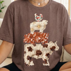 May include: Brown t-shirt featuring a stacked design of farm animals. The animals include a cow, pig, sheep, and chicken, each with unique floral patterns. The cow is at the bottom, with the pig, sheep, and chicken stacked on top.
