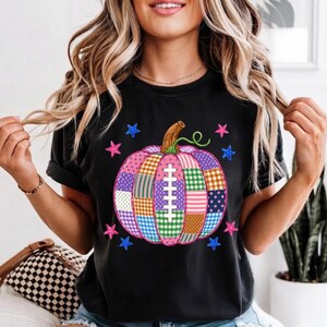 May include: A black t-shirt featuring a large, central pumpkin graphic composed of various colourful plaid and polka-dot patches. The pumpkin includes a white football lace design in the centre and is surrounded by small pink and blue stars.