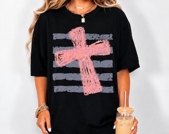 May include: A black short-sleeve t-shirt featuring a large, distressed-style pink cross graphic centered over a series of horizontal, weathered grey stripes. The shirt has a crew neckline and a relaxed fit, showcasing a bold, textured design.