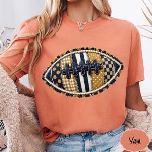Puede incluir: Una camiseta de color coral con un gráfico central de un balón de fútbol americano. El diseño del balón se compone de varios parches estampados en azul marino, dorado y blanco, incluyendo lunares, rayas y cuadros vichy, con un borde de cuentas.