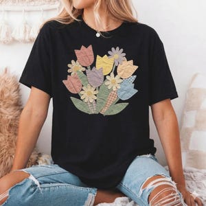 May include: Black t-shirt with a floral design featuring tulips, daisies, and leaves in pastel colours and plaid patterns. The shirt is a casual, short-sleeved style.