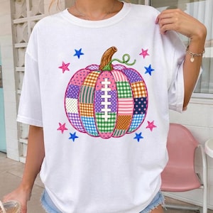 May include: A white short-sleeved t-shirt featuring a large, central graphic of a pumpkin made of various colorful patchwork patterns, including plaid, polka dots, and stripes, with a football lace design in the center and small stars.