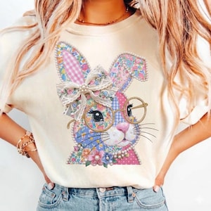 May include: Cream-colored t-shirt featuring a colorful Easter bunny design. The bunny has floral patterned ears, a bow, and round glasses. The design includes pink, blue, and white colors, with a pearl necklace and floral accents.
