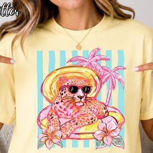 Puede incluir: Camiseta amarilla clara con un diseño de leopardo con gafas de sol y sombrero, descansando en un flotador. El diseño incluye palmeras y flores rosas, con rayas azules y blancas. La palabra "Butter" está en la esquina superior izquierda.