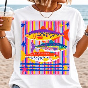 Puede incluir: Una camiseta blanca con un estampado gráfico de tres peces estilizados en amarillo, morado, azul y naranja, sobre un fondo de rayas verticales y horizontales multicolores y pequeñas estrellas en rosa, azul y amarillo.