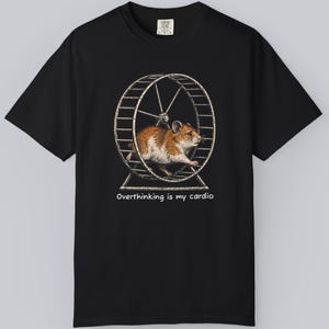 May include: A black short-sleeve t-shirt featuring a graphic of a brown and white hamster running inside a metal exercise wheel. Below the graphic, the text reads, "Overthinking is my cardio." The shirt has a standard crew neckline and short sleeves.
