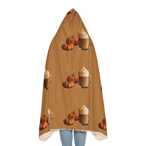 Puede incluir: Una manta con capucha con un patr&oacute;n repetido de calabazas y bebidas de caf&eacute;. La manta es principalmente marr&oacute;n con un interior color crema. Las calabazas son naranjas y las bebidas de caf&eacute; tienen crema batida.