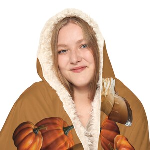 Puede incluir: Una manta con capucha con tem&aacute;tica oto&ntilde;al, con calabazas naranjas y una bebida con crema batida. La manta tiene un exterior marr&oacute;n y un forro interior cremoso y esponjoso alrededor de la capucha.