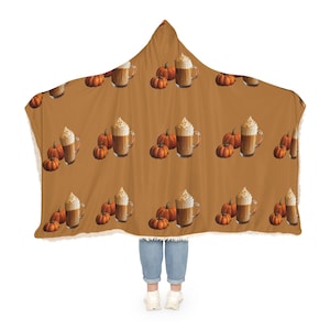 Puede incluir: Una manta con capucha con un patr&oacute;n repetido de calabazas y lattes de especias de calabaza sobre un fondo marr&oacute;n. La manta tiene un ribete blanco y esponjoso. La persona que lleva la manta lleva vaqueros azules y zapatillas blancas.