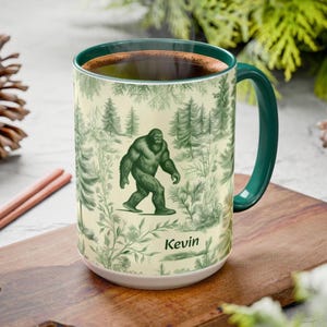 May include: A large ceramic mug with a green handle and rim, featuring a forest scene with a green Bigfoot illustration. The mug has the name "Kevin" printed on it. The mug is filled with a dark liquid.