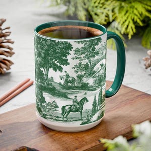 Puede incluir: Una taza de cerámica blanca con asa y borde verdes, llena de café. La taza presenta un diseño toile de Jouy verde que representa jinetes a caballo en un entorno rural. La taza está sobre una tabla de cortar de madera.