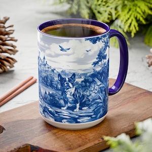 May include: A blue and white ceramic coffee mug with a purple handle. The mug features a blue and white illustration of a French bulldog standing on a rock in a forest setting. The mug is filled with coffee and is sitting on a wooden cutting board.