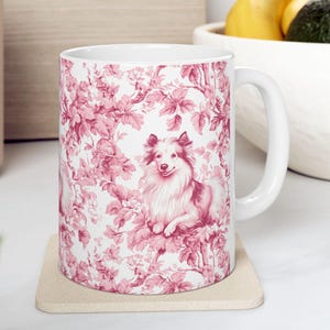 Puede incluir: Taza de cerámica blanca con un diseño toile de Jouy rosa y blanco, que representa un perro entre elementos florales. La taza tiene un asa curva y está sobre un posavasos beige. El diseño es clásico.