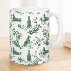 May include: White ceramic mug featuring a green dog and floral toile pattern. The mug has a curved handle and is set on a wooden surface. The design includes various poses of the dog, surrounded by foliage.