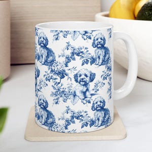 May include: White ceramic mug with a blue and white floral pattern featuring a repeating image of a small dog. The mug has a handle and is sitting on a beige coaster.