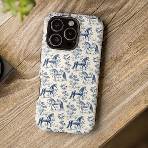Könnte beinhalten: Eine Handyhülle mit einem blau-weißen Toile-de-Jouy-Muster mit Pferden und floralen Elementen. Die Hülle liegt auf einer Holzoberfläche. Die Hülle hat eine glänzende Oberfläche und ein schützendes Design.
