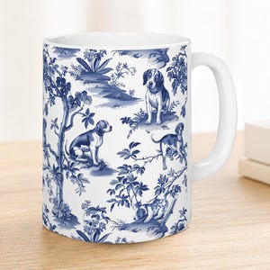 May include: White ceramic mug with a blue and white toile pattern depicting dogs, trees, and plants. The mug has a curved handle and is on a wooden surface. A vase with white flowers is to the left.