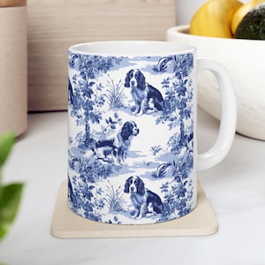 May include: White ceramic mug with a blue and white pattern of dogs and flowers. The mug has a handle and is sitting on a beige coaster.