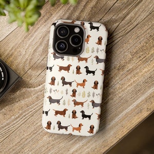 Puede incluir: Una funda blanca para iPhone con un patrón de dachshunds marrones, negros y tostados. La funda tiene un recorte para el objetivo de la cámara y un fondo blanco.