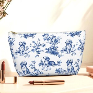 May include: White cosmetic bag with a blue toile pattern featuring dachshund dogs and floral elements. The bag has a gold zipper and is sitting on a light-colored surface. Lipstick and a lip pencil are also on the surface.