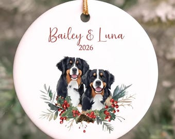 May include: A round white ceramic ornament hanging from a gold string. The text Bailey & Luna 2026 is printed in red. Below the text are two Bernese Mountain Dogs surrounded by a festive illustration of green pine branches and red berries.