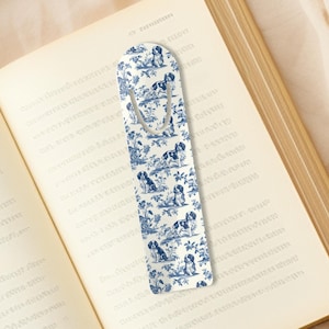 May include: A white bookmark with a blue and white pattern of dogs and flowers. The bookmark has a metal clip at the top.