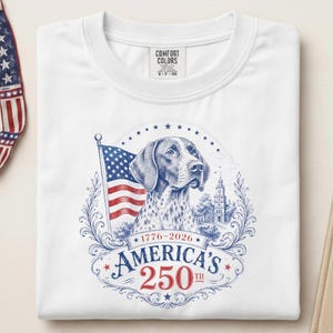 May include: A white t-shirt featuring a graphic print of a dog, an American flag, and a building. The text reads 1776-2026 AMERICA'S 250TH. The design is framed by blue and red decorative scrollwork and stars, with a Comfort Colors brand label inside.