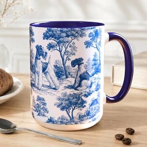 Może przedstawiać: Biały ceramiczny kubek z niebiesko-białym wzorem toile de Jouy przedstawiającym dwa psy w krajobrazie. Kubek ma ciemnoniebieskie wnętrze i uchwyt. Rogalik, łyżka i ziarna kawy leżą na drewnianym stole.