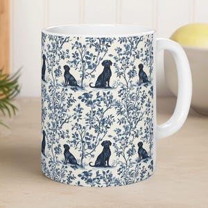May include: White ceramic mug with a blue floral and black dog design. The mug has a white handle and a smooth surface. The pattern is repeated around the mug. The design is reminiscent of a classic toile.