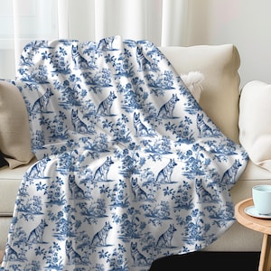 May include: A white blanket with a blue toile pattern featuring German Shepherd dogs and floral elements. The blanket is draped over a beige sofa, creating a cozy and decorative look. The design is repeated across the blanket.