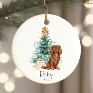 May include: White ceramic Christmas ornament featuring a brown Cavalier King Charles Spaniel, a teal Christmas tree, and wrapped gifts. The ornament has a gold ribbon for hanging and the name "Ruby" and the year "2025" are printed on the front.