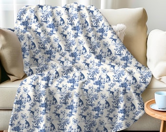 May include: A white and blue throw blanket with a repeating pattern of blue dogs in a floral design. The blanket is draped over a white sofa.