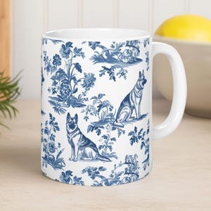 May include: White ceramic mug with a blue and white toile pattern featuring German Shepherd dogs and floral designs. The mug has a white handle and is suitable for hot or cold beverages.