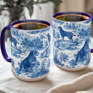 May include: Two ceramic mugs with a blue and white toile design featuring dogs in a landscape. The mugs have purple handles and rims, and are filled with a dark liquid. The mugs are sitting on a white surface.
